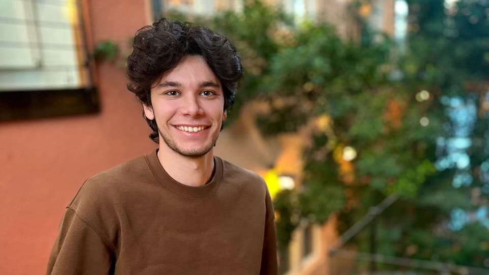 A young man with black curly hair and a brown sweater. In the background, green vegetation and stone walls in red and beige can be seen. 